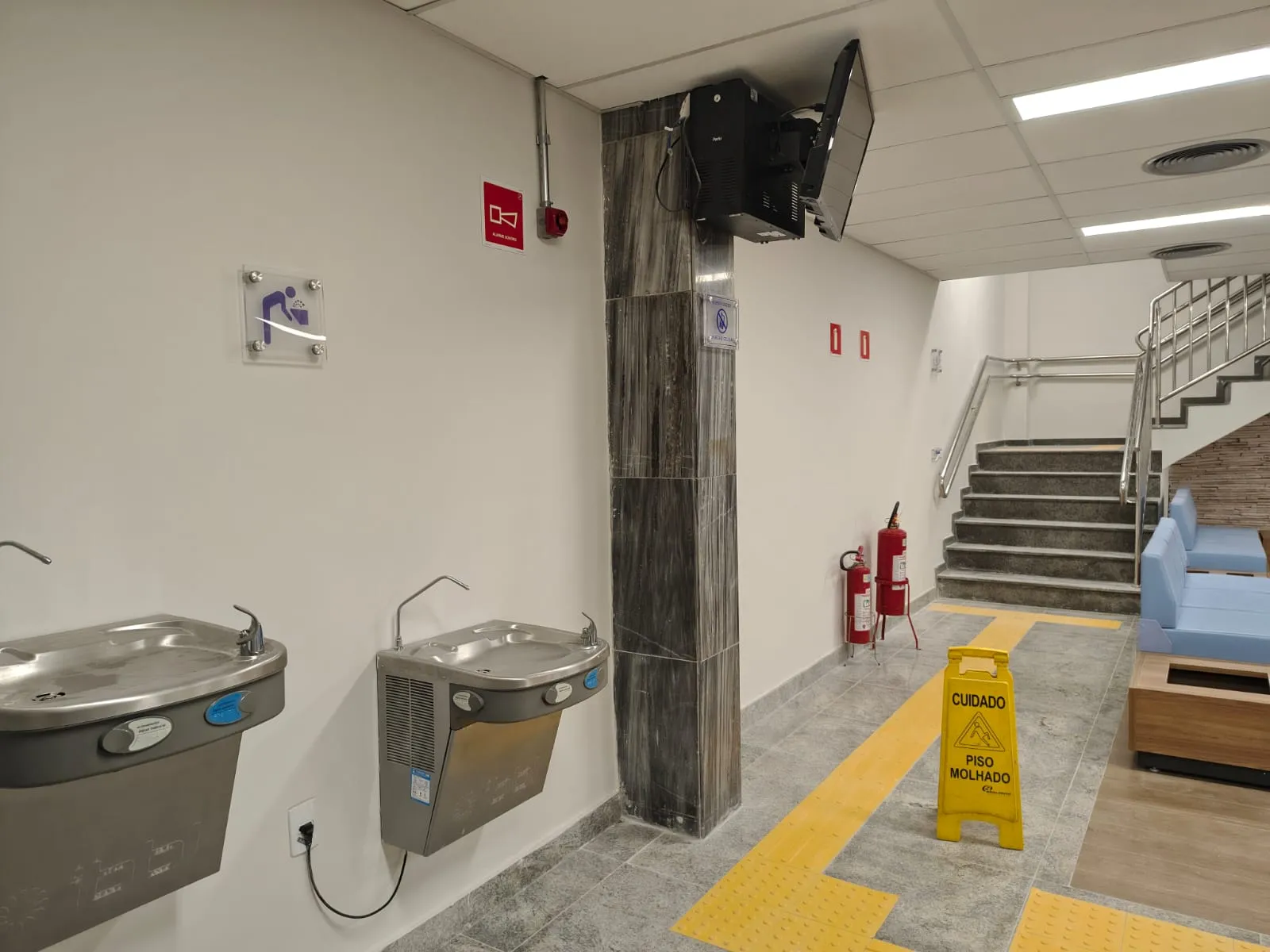 Sala de espera com bebedouros, sofás azuis e escada com sinalização